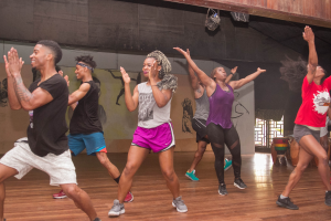 Gruppe von Menschen, die in einem Studio mit einer bemalten Wand, einer Glasscheibe und Deckenbeleuchtung tanzen, wahrscheinlich an einem Zumba-Kurs teilnehmen.