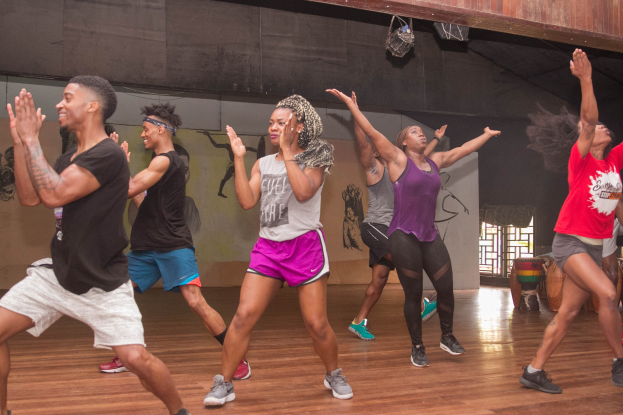 Gruppe von Menschen, die in einem Studio mit einer bemalten Wand, einer Glasscheibe und Deckenbeleuchtung tanzen, wahrscheinlich an einem Zumba-Kurs teilnehmen.