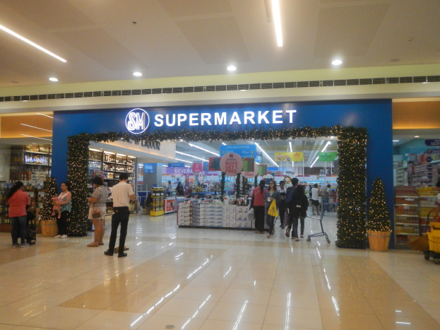 Eine Gruppe von Menschen, die vor einem Supermarkt stehen, einige halten Einkaufstaschen, mit einem Einkaufswagen, Topfpflanze, Regalen, Schildern und Deckenbeleuchtung im Hintergrund.