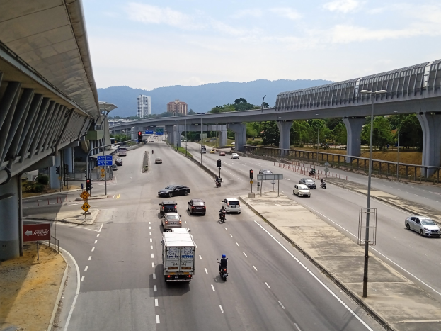 Eine stark befahrene Autobahn verläuft parallel zu einer Brücke, flankiert von Straßenlaternen, Ampeln, Schildern, Bäumen, Gebäuden und Hügeln unter einem bewölkten Himmel.