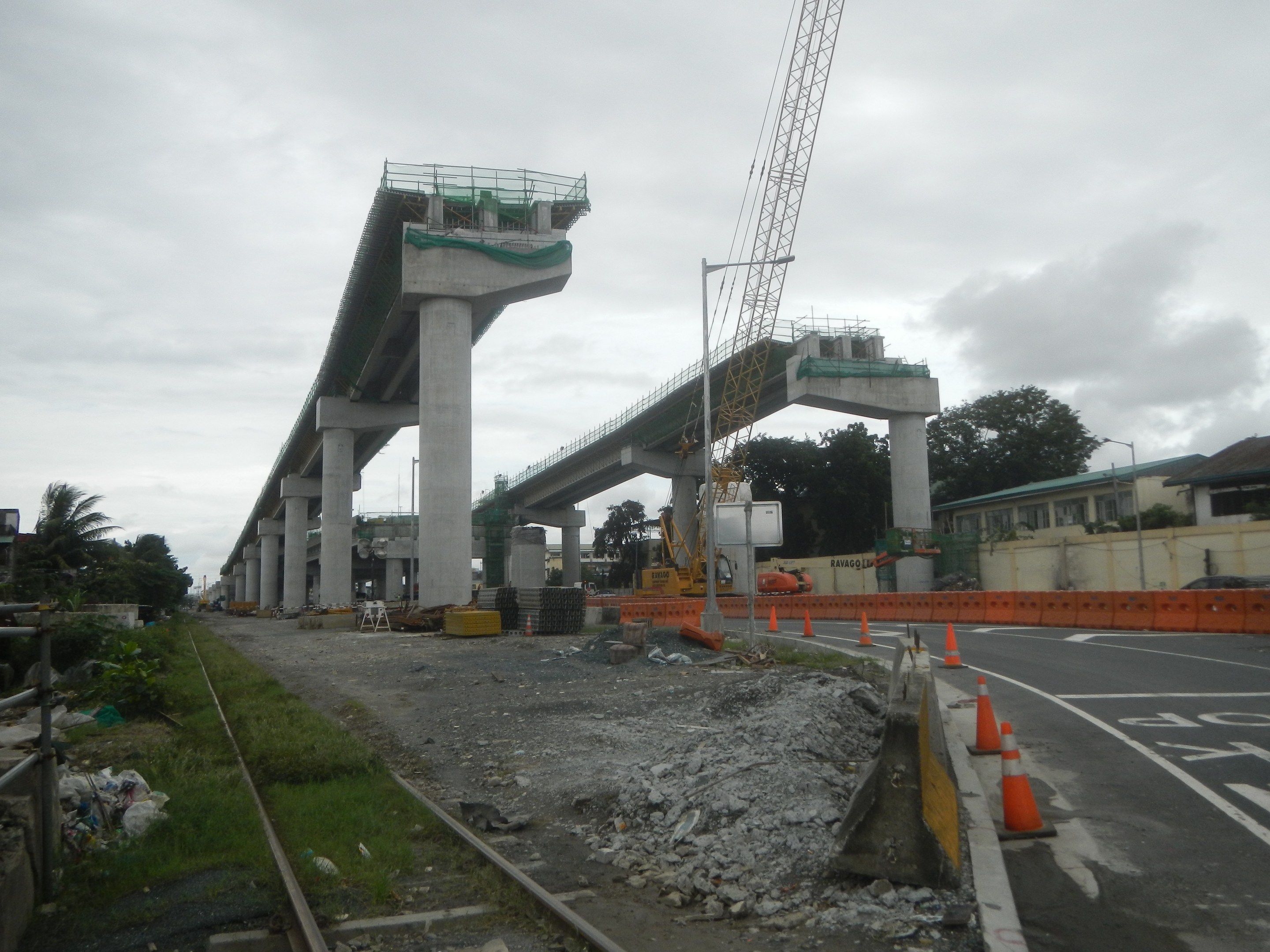 Baustelle mit einer Brücke im Hintergrund, Straße mit Absperrkegeln markiert, Bahnschiene links, verstreute Steine und Gras, Bäume und Gebäude auf beiden Seiten und ein bewölkter Himmel.