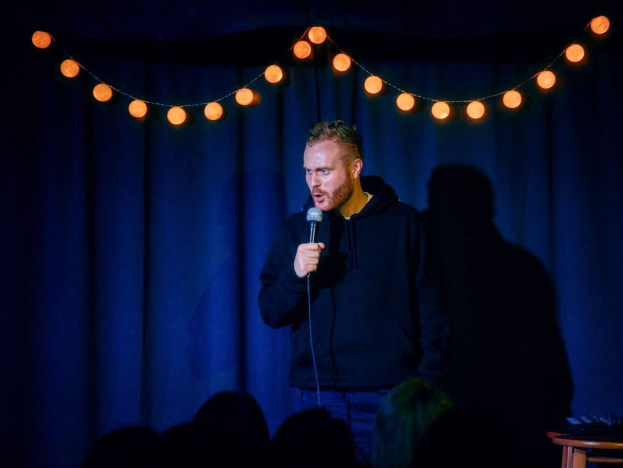 Ein Mann hält ein Mikrofon in der Hand und steht auf einer hell erleuchteten Bühne bei einem Stand-up-Comedy-Event, rechts neben ihm steht ein Tisch mit verschiedenen Gegenständen und im Hintergrund ist das Publikum zu sehen.