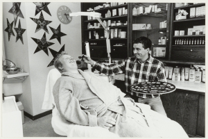 Ein Mann lächelt in einem Zahnarztstuhl, während ein anderer Mann neben ihm steht und ein Tablett hält, mit einem Waschbecken, einer Uhr an der Wand und Regalen voller Flaschen im Hintergrund.