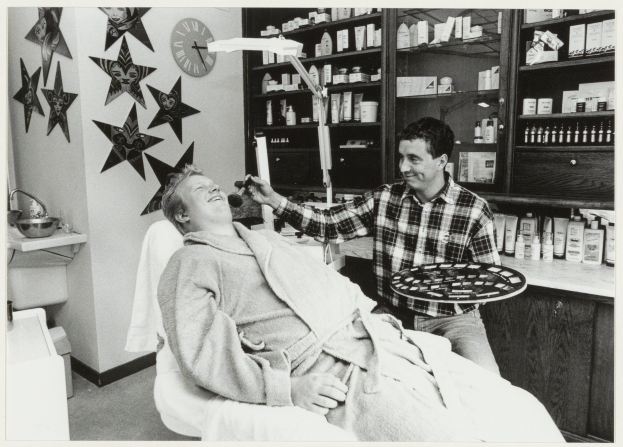 Ein Mann lächelt in einem Zahnarztstuhl, während ein anderer Mann neben ihm steht und ein Tablett hält, mit einem Waschbecken, einer Uhr an der Wand und Regalen voller Flaschen im Hintergrund.