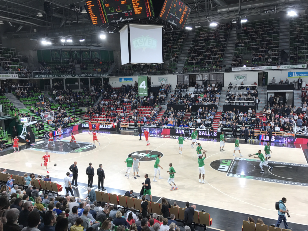 Basketballspiel in einer großen Arena mit Zuschauern auf den Rängen, Anzeigetafeln und beleuchtetem Spielfeld.