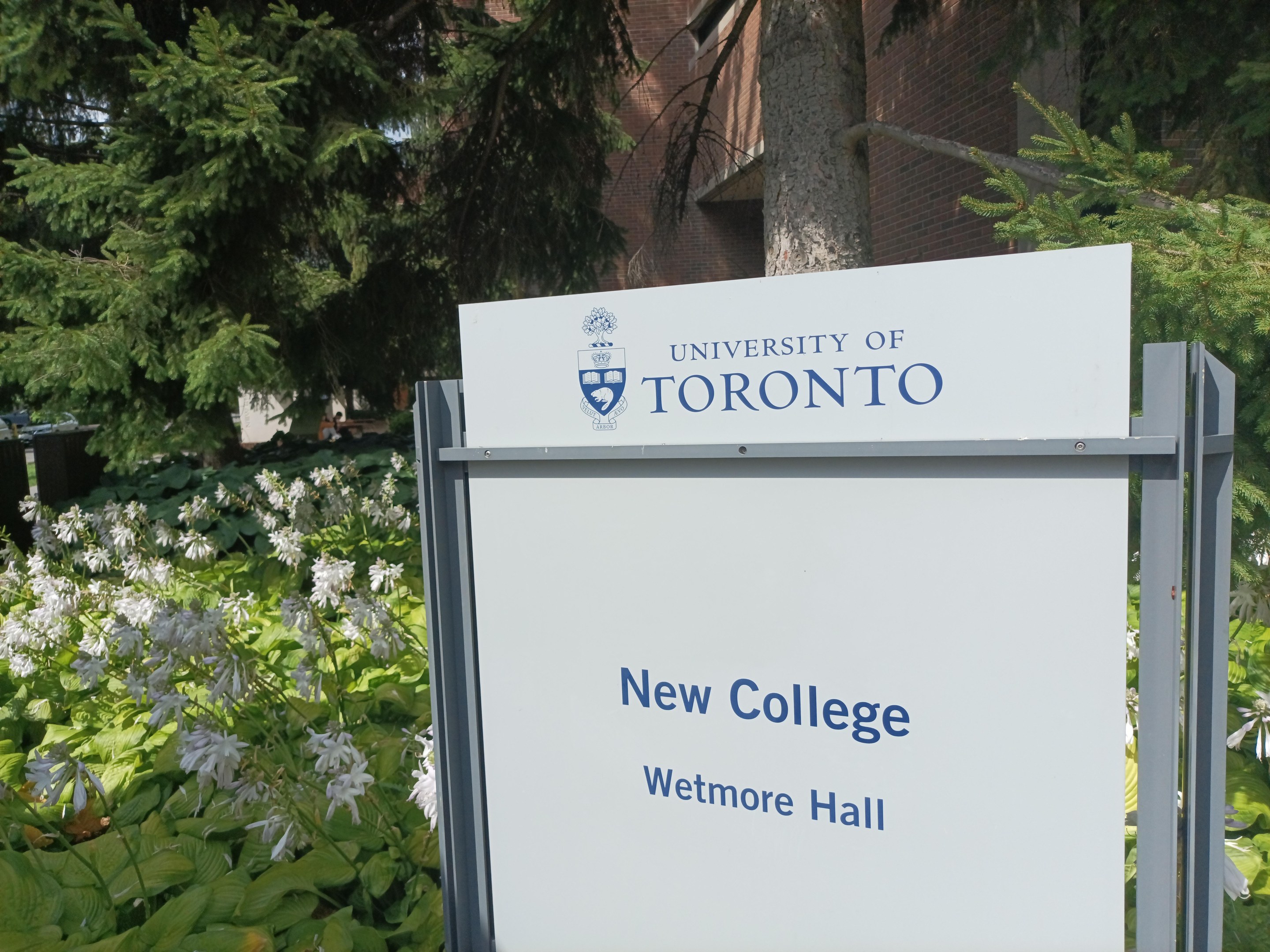 Außenansicht des neuen Colleges der Universität Toronto an der Wetmore Hall, mit einem Schild vor dem Gebäude, umgeben von grünem Gras, bunten Blumen, hohen Bäumen und einem klaren blauen Himmel.