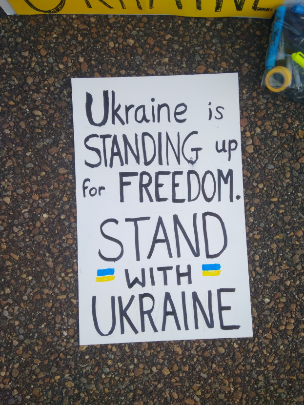 Ein weißer Papierbogen mit den Worten "Ukraine steht für Freiheit, stehen Sie mit Ukraine" liegt vor einer gelben Fahne auf dem Boden, rechts daneben eine Plastikabdeckung mit verschiedenen Gegenständen.