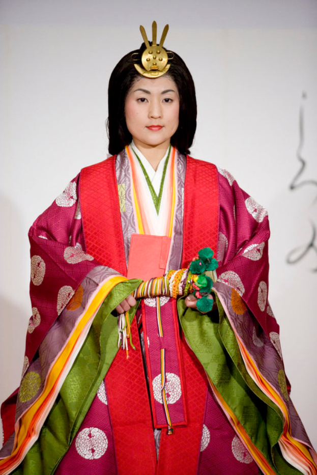 Eine Frau in einem traditionellen japanischen Kimono, das ein leuchtend rotes und grünes Kleid mit kunstvollen Mustern trägt, steht selbstbewusst auf einem Laufsteg, ihr Haar ist ordentlich zurückgebunden.