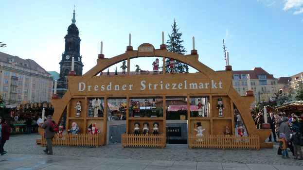 Ein geschäftiger Weihnachtsmarkt in Dresden, Deutschland, mit Menschen, die gehen, einige mit Taschen, unter einem Bogen, mit Bäumen und Gebäuden im Hintergrund unter einem bewölkten Himmel.