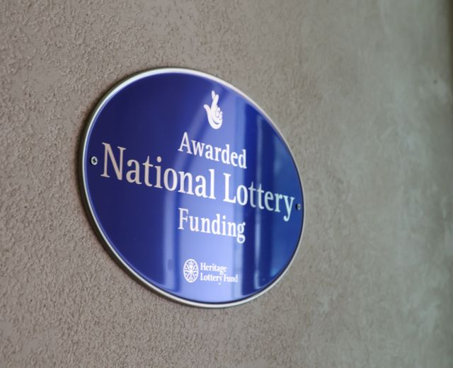 Blaues rechteckiges Schild an einem Gebäude mit weißem Hintergrund und schwarzer fetter Schrift, die "National Lottery Funding awarded" lautet.