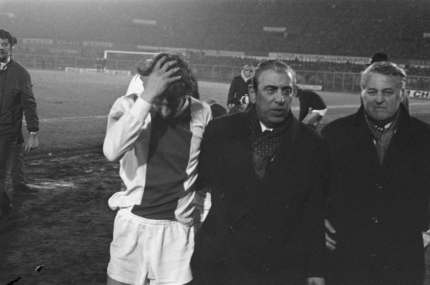 Schwarzes und weißes Bild einer Gruppe von Männern auf einem Fußballfeld mit einem Stadion im Hintergrund, die enttäuscht nach einem Matchsieg aussehen.