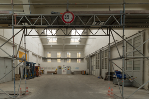 Ein großes Lager mit ausgedehnten Gerüsten, Verkehrskegeln auf dem Boden, Metallstangen, ein Schild, verschiedene Maschinen, eine Tür, Fenster, eine Wand und ein Dach mit Deckenleuchten.