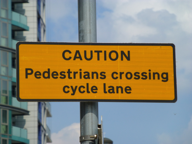 Gelbes Warnschild auf einem Pfosten mit der Aufschrift "Fußgänger überqueren Radweg" vor einem Gebäude unter einem klaren blauen Himmel.