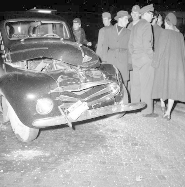 Schwarz-weiß-Bild eines schwer beschädigten Autos am Straßenrand mit eingedellter Motorhaube und zersplitterten Fenstern, umgeben von einer Gruppe von Menschen, die die Szene begutachten.