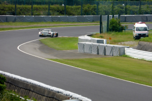 Ein Rennwagen fährt auf einer Rennstrecke mit einem im Hintergrund sichtbaren Polizeiwagen, umgeben von Gras, Pflanzen, Bäumen und Absperrungen, mit einem Fahrzeug auf der rechten Seite und Text unten.