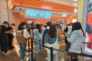 Eine Gruppe von Menschen steht an einem Tresen in einem Restaurant Schlange, einige tragen Kappen und Taschen, andere halten Gegenstände, mit Stangen mit Bändern, einem Bildschirm mit Text, Deckenleuchten und Tischen mit verschiedenen Gegenständen im Hintergrund.