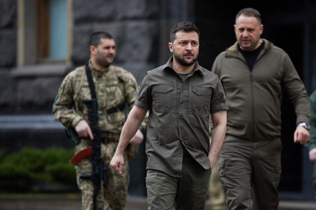 Ein Mann in militärischer Uniform, identifiziert als ehemaliger ukrainischer Präsident Viktor Janukowytsch, wird von zwei bewaffneten Männern in militärischer Uniform vor einem Gebäude mit Pflanzen im Hintergrund eskortiert.