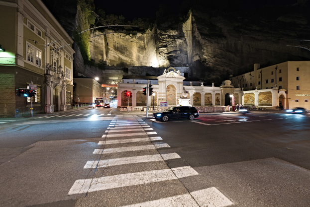 Nachtsichtige Stadtstraße mit fahrenden Autos, beleuchtet von Straßenlaternen und Ampeln, Gebäude mit beleuchteten Fenstern im Hintergrund, ein Hügel, Laternenmasten und eine Zebrastreifenüberquerung unten.