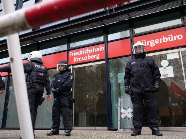 Eine Gruppe von Polizisten in Helmen und Handschuhen steht vor einem Gebäude mit Glaswänden und -türen, neben einem Pfahl, mit "Friedrichshain Kreuzberg" auf Schildern im Hintergrund und Papieren an der Glaswand.