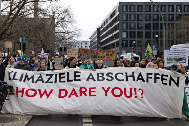 Gruppe von Menschen, die eine Straße entlanggehen und ein Banner mit der Aufschrift "Klimaziele Abschaffen - Wie Wagst Du Es?" halten, mit Fahrrädern auf der linken Seite und Gebäuden, Bäumen und einem klaren blauen Himmel im Hintergrund.