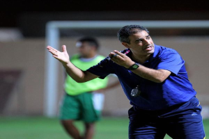 Ein Mann in einem blauen T-Shirt und Hose steht auf einem Fußballfeld und hält ein Mikrofon in der Hand, während er auf Spieler zeigt. Im Hintergrund ist eine Person in einem grünen Kleid, ein Torpfosten und eine Wand zu sehen.