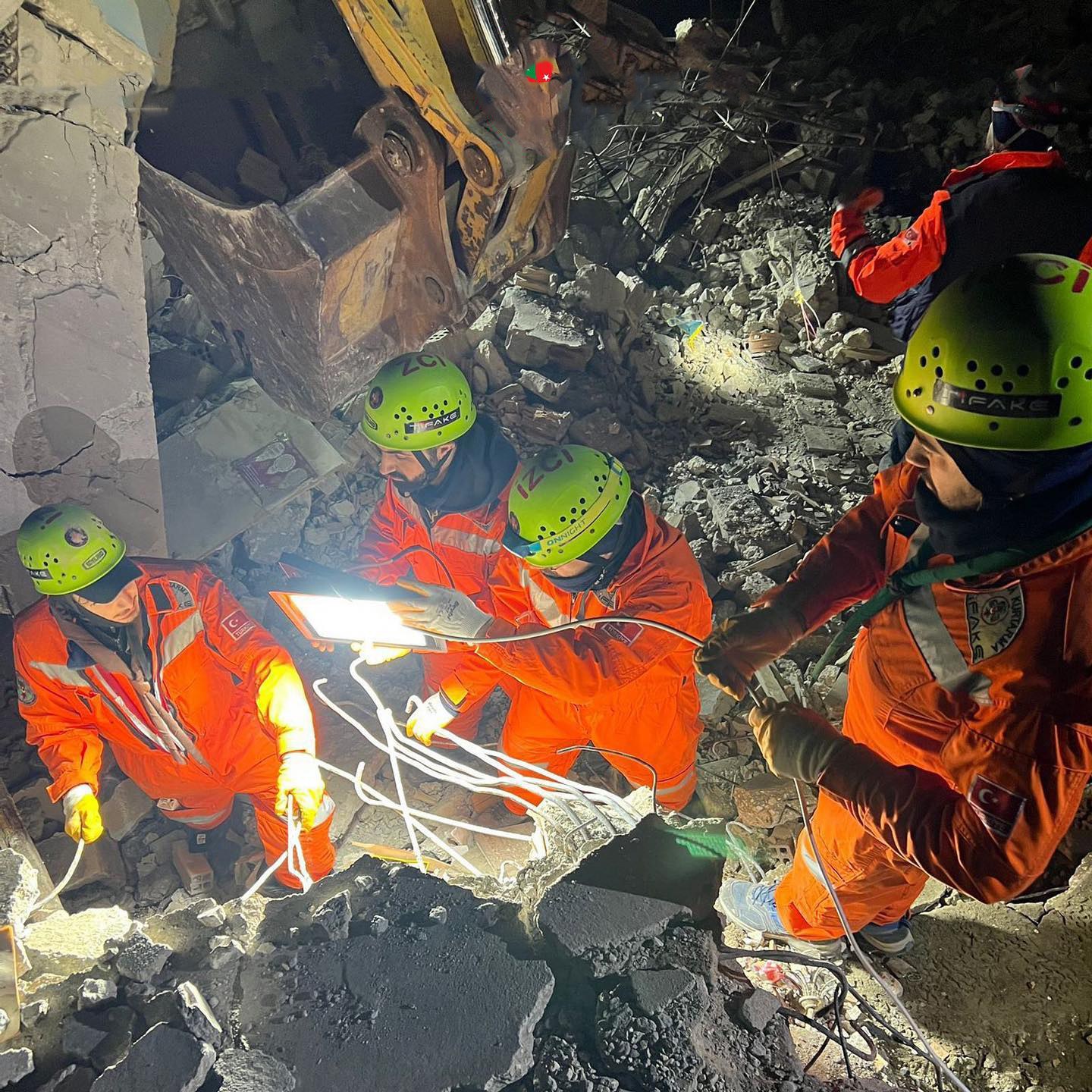 Männer in orange Uniformen und grünen Helmen bei der Arbeit an Schutt mit einem Kran im Hintergrund bei einer Rettungsaktion nach einem Einsturz.