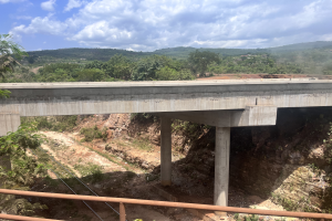 Eine Brücke über einen Schotterweg mit Bäumen, Hügeln und einem bewölkten Himmel im Hintergrund sowie einem Geländer am Boden, das auf ein laufendes Bauprojekt hinweist.