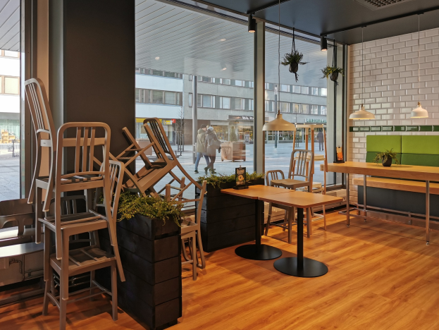 Ein Restaurant in Berlin, Deutschland, mit hölzernen Tischen und Stühlen in der Nähe eines großen Fensters, das Menschen zeigt, die auf der Straße gehen, Gebäude und den Himmel umgeben von Pflanzen und Lichtern.