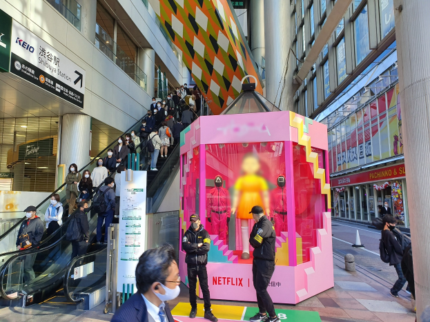 Eine Gruppe von Menschen steht vor einem Glaswand-Gebäude mit Säulen und Texttafeln, einige tragen Masken, während eine Rolltreppe mit Menschen im Hintergrund fährt; ein pinker Kasten in der Mitte zeigt Schaufensterpuppen und trägt die Aufschrift "Netflix's neuer Netflix-Stand in Tokyo."