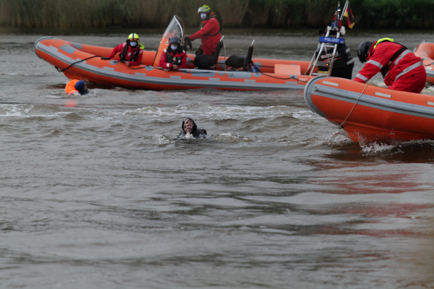 Eine Gruppe von Menschen in einem aufblasbaren Boot auf einem Fluss, mit zwei Personen im Wasser im Vordergrund und Vegetation im Hintergrund, alle mit Schwimmwesten und Helmen ausgestattet, was auf einen Rettungseinsatz hinweist.