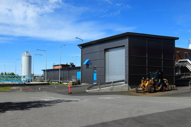 Großes Industriegebäude mit einem Bagger davor, umgeben von Straßeninfrastruktur und Fahrzeugen unter einem bewölkten Himmel.