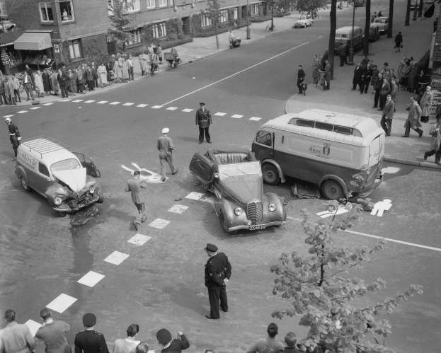 Schwarz-weiß-Szene eines Autounfalls an einer Straßenkreuzung mit mehreren Fahrzeugen, darunter ein Lieferwagen, und einer Gruppe von Menschen in der Nähe, mit Gebäuden, Bäumen, Pfosten und Texttafeln im Hintergrund.