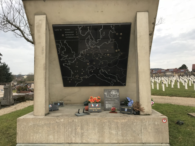 Ein Friedhofdenkmal mit einer Karte, umgeben von Blumen und Gegenständen, das das Grab eines deutschen Weltkriegsoldaten markiert, mit Gräbern, Bäumen, Gebäuden und einem blauen Himmel im Hintergrund.