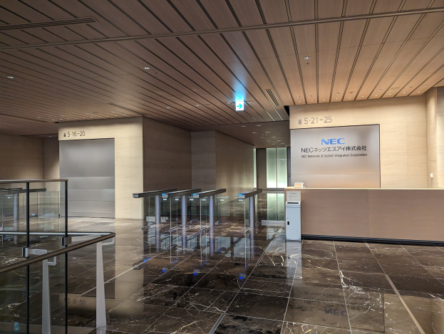 Eingangshalle eines Gebäudes mit Marmorboden, Glasgeländern, elektrischen Beleuchtungen bis zur Decke, Namensschildern an den Wänden und einem Empfangstisch sowie einem Logo, das auf die NEC Network & System Integration Corporation hinweist.