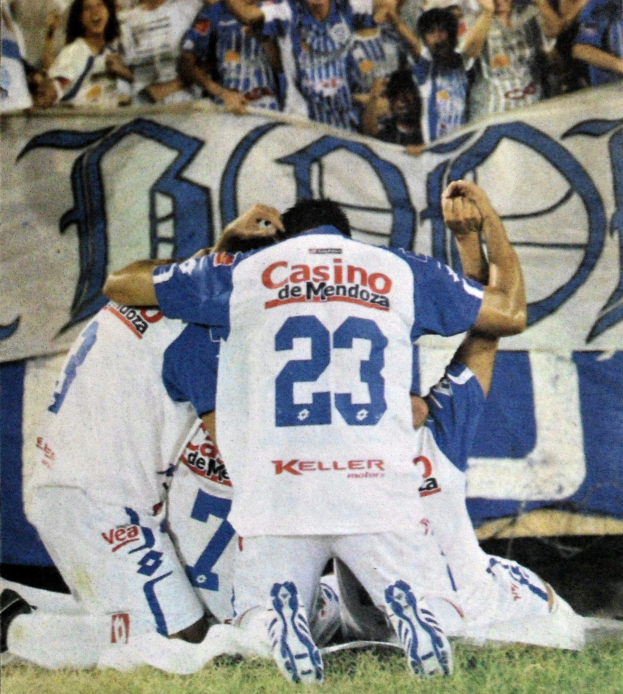 Gruppe von Fußballspielern feiert vor jubelnder Menge auf dem Boden, mit einem Banner mit Text im Hintergrund.