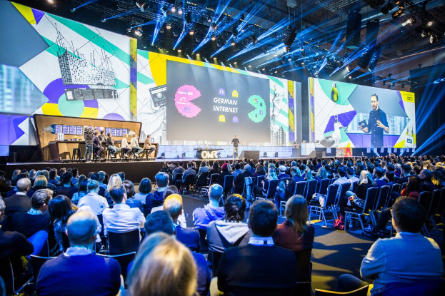 Eine große Menschenmenge sitzt auf Stühlen, die einer Bühne zugewandt sind, mit einem Tisch voller Gegenstände auf der linken Seite und Bildschirmen und Lichtern im Hintergrund.