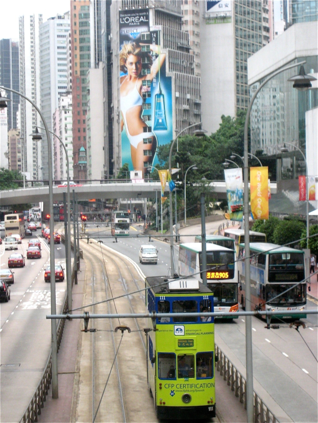 Eine belebte Stadtstraße mit verschiedenen Fahrzeugen, darunter Doppeldeckerbusse, Autos und andere Fahrzeuge, unter einem klaren blauen Himmel, umgeben von hohen Gebäuden, Polen, Lampen, Tafeln, Bannern, Bäumen und einer Brücke, mit Text am unteren Bildrand.