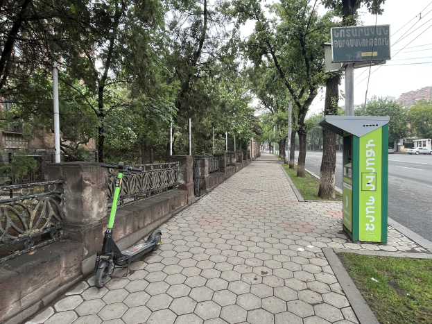 Elektrisches Scooter neben einem Bushaltestellen-Schild auf einer Stra├če mit fahrenden Fahrzeugen, Gras, B├Ąumen, Strommasten, Geb├Ąuden und einem Müll├Ęimer im Hintergrund.