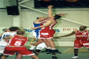 Eine Gruppe von Männern, die Basketball auf einem Platz mit einer Wand im Hintergrund spielen, auf der Text und Lautsprecher zu sehen sind, und ein Basketball in der Luft oben auf dem Bild.
