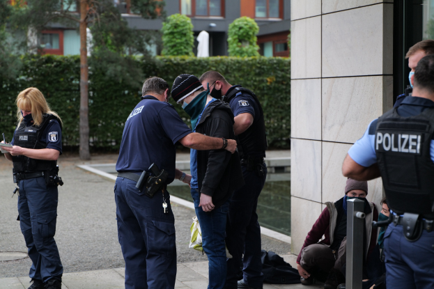 Gruppe von Polizisten vor einem Gebäude mit einer Frau, die ein Buch und einen Stift hält, umgeben von sitzenden und stehenden Personen, mit Bäumen und Gebäuden im Hintergrund.