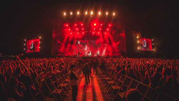 Eine große Menschenmenge steht vor einer hell erleuchteten Bühne mit Geländern, auf der Musiker spielen, während andere zusehen; der Hintergrund ist dunkel.
