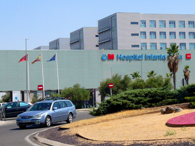 Ein Krankenhausgebäude mit parkenden Autos, umgeben von städtischer Infrastruktur und Grünflächen unter einem klaren Himmel.