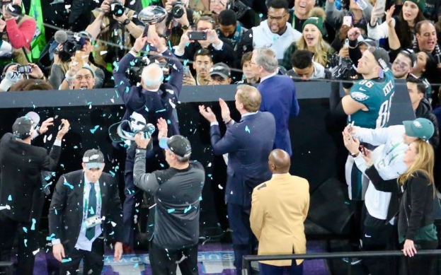 Philadelphia Eagles Spieler feiern mit dem Vince Lombardi Trophy nach dem Gewinn des Super Bowls, tragen Mützen und werfen Konfetti, mit einem Geländer und Fotografen im Hintergrund.