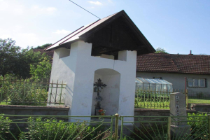 Kleine weiße Kirche mit einem Kreuz darauf, umgeben von einem Metallzaun, Pflanzen, Gras, einem Weg, Häusern, Polen, Drähten und einem bewölkten Himmel, identifiziert als die Kirche des Heiligen Grabes.