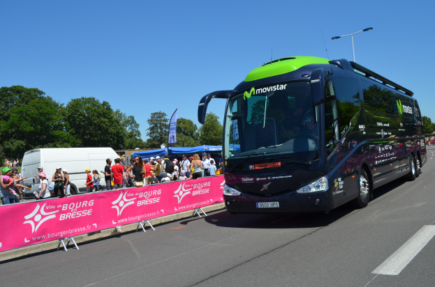 Ein schwarzer und grüner Bus fährt auf einer Straße neben einer Menschenmenge, einige tragen Möppen, mit einem Banner auf der linken Seite und Bäumen unter einem klaren blauen Himmel im Hintergrund.