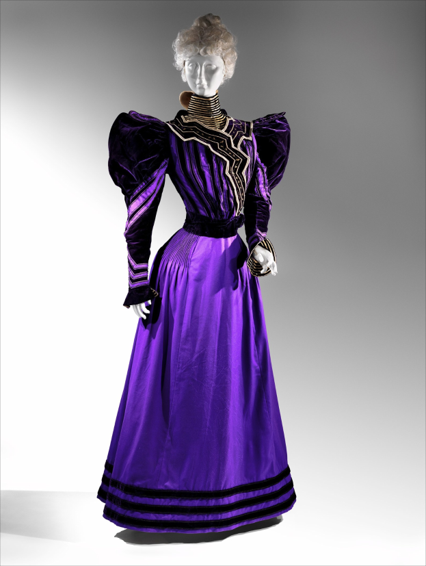 Eine Schaufensterpuppe in einem bodenlangen lila Abendkleid mit einem schwarzen und goldenen gemusterten Rock, der goldene Stickerei und Perlen enthält, einem eng anliegenden Oberteil mit Spitzenbesatz und frisierten Haaren mit zarten Ohrringen.