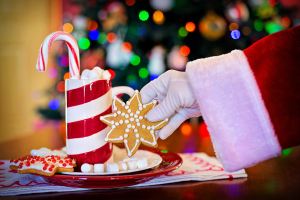 Person in einem Santa Claus Hut, die eine Tasse heißen Schokolade und einen Zuckerstange hält, mit einem Teller mit Keksen und einem Tuch auf dem Tisch vor ihnen, vor einem Weihnachtsbaum und einer Wand.