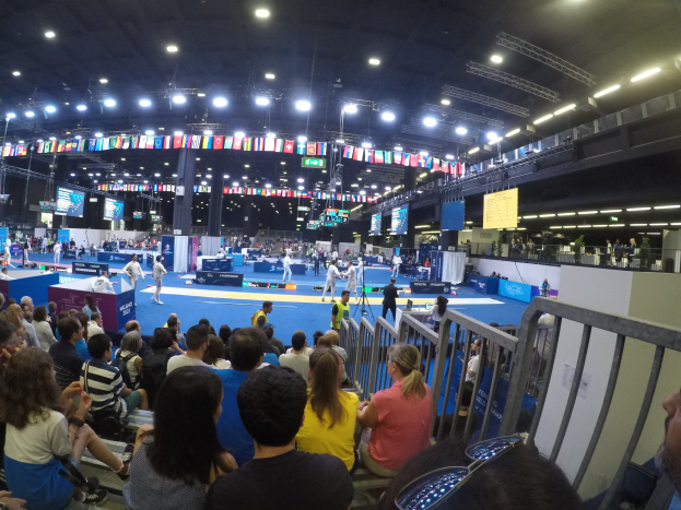Gro├če Menge sitzend und stehend in einer Indoor-Sportarena mit Bildschirmen, Fahnen und Deckenbeleuchtung im Hintergrund.