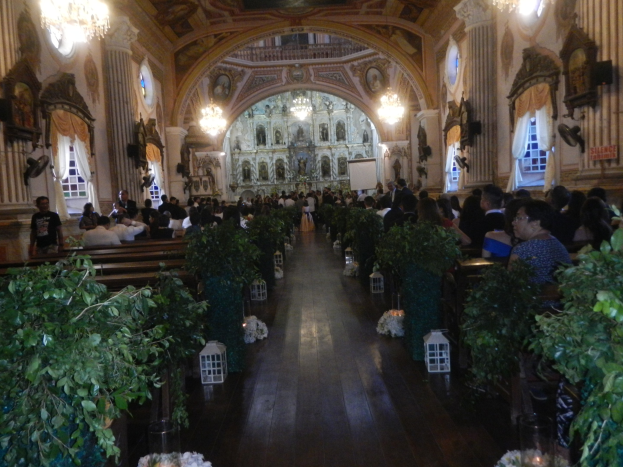 Eine Hochzeitszeremonie in einer Kirche mit Gästen auf Bänken sitzend und im Gang stehend, dekoriert mit Zimmerpflanzen, Skulpturen an den Wänden, Vorhängen an den Fenstern und hängenden Kronleuchtern, mit einem Bildschirm im Hintergrund.