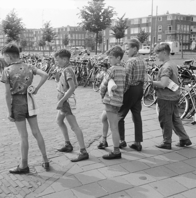 Schwarzes und weißes Foto einer Gruppe junger Jungs, die auf einem Bürgersteig stehen, umgeben von Fahrrädern, Fahrzeugen, Bäumen, Pfählen und Gebäuden.